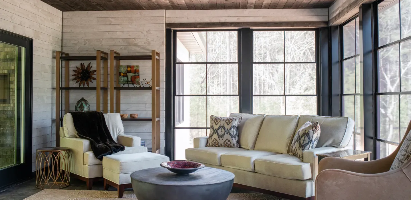 Bright sunroom with beige sofa, armchair, round coffee table, and large windows overlooking trees.