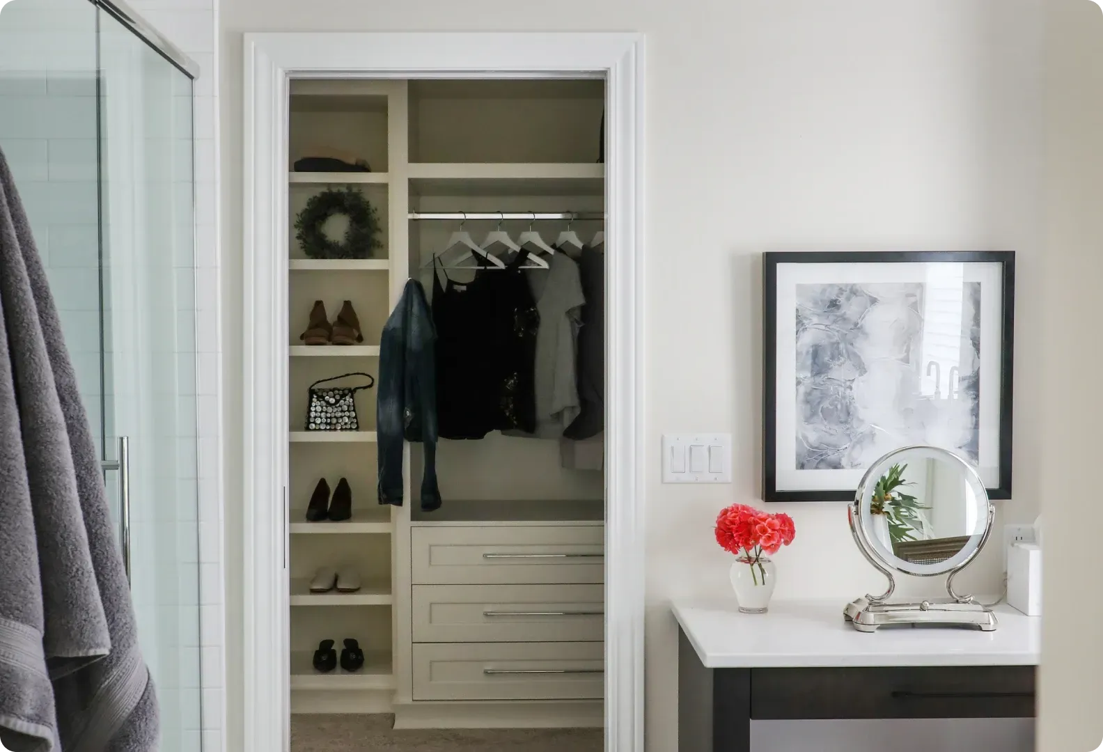 Open closet with clothes and shelves beside a vanity table with mirror and red flowers