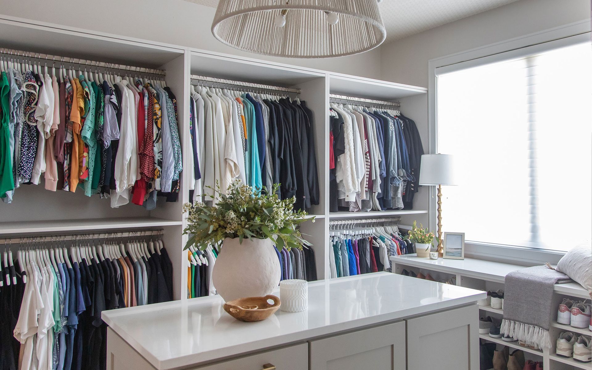 Bright closet room with hanging clothes, a white island, and a plant near a sunlit window.