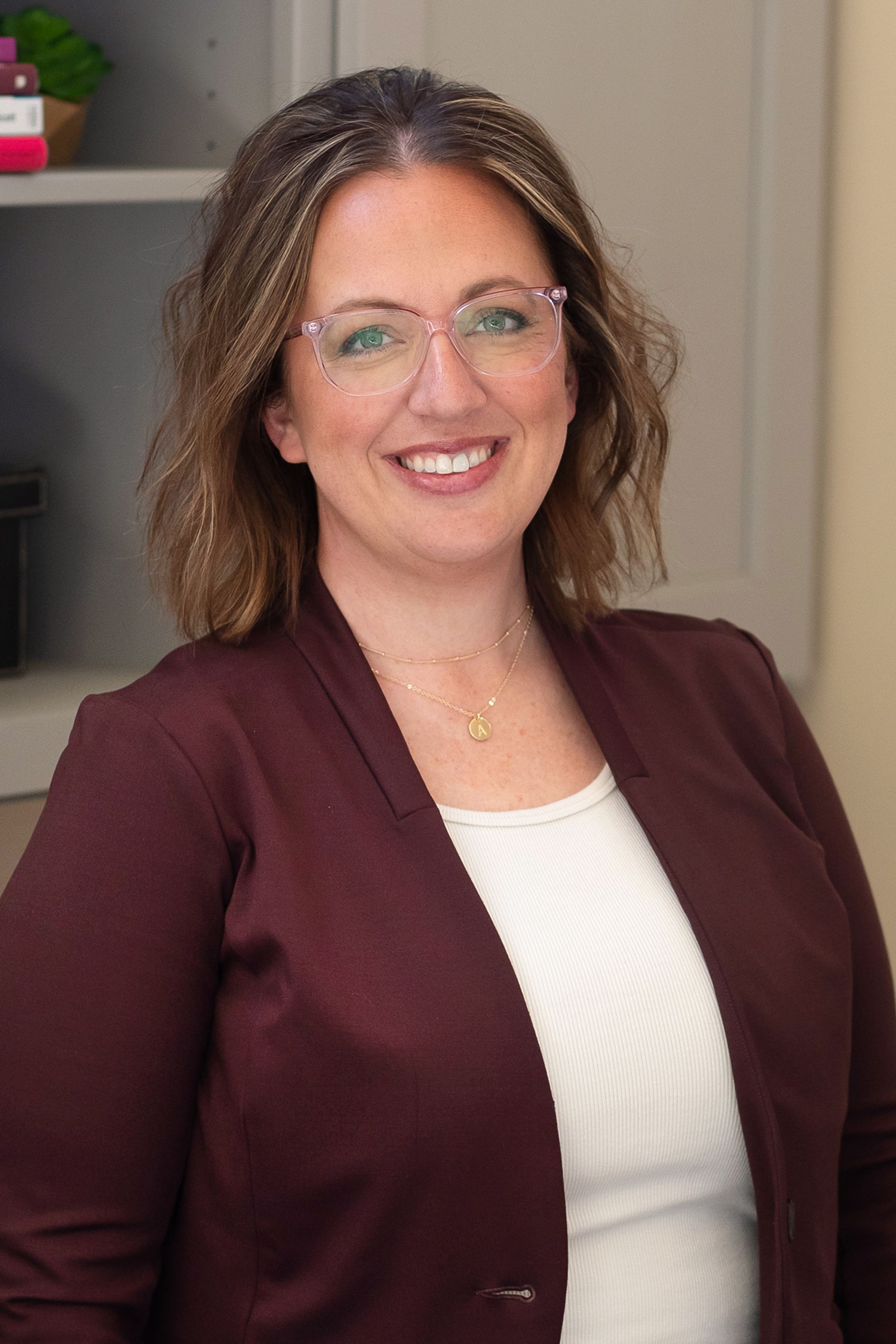 Smiling woman with glasses in a maroon blazer, standing indoors in a home setting