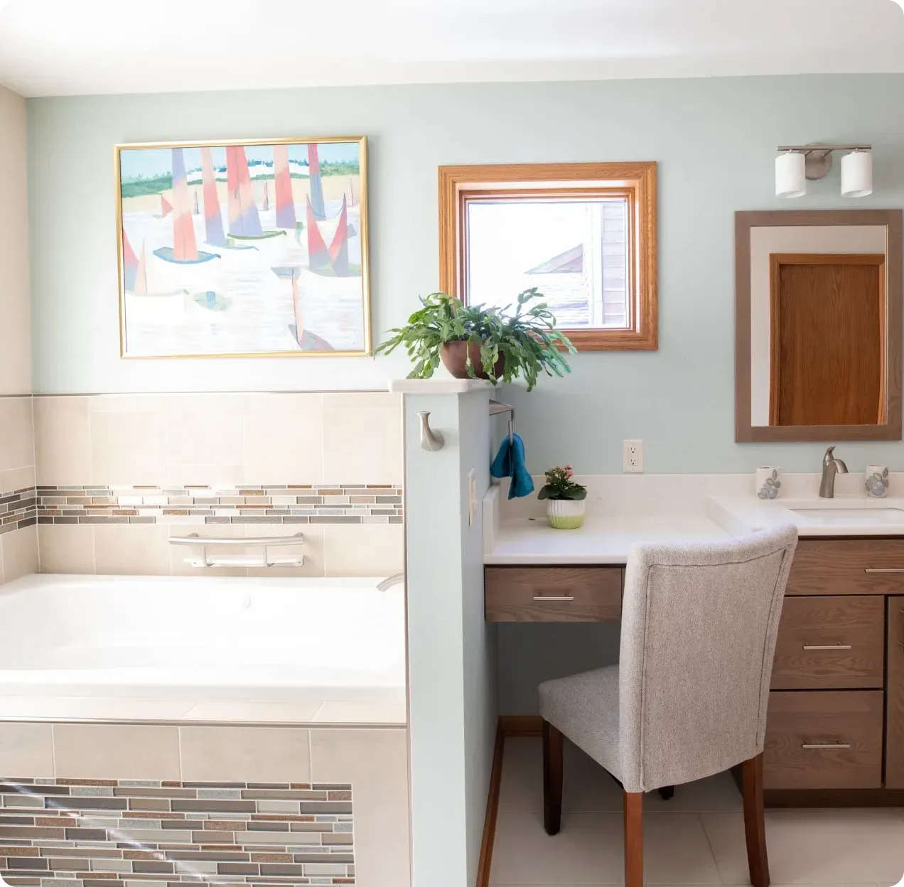 Bright bathroom vanity with chair, mirror, flowers, and a bathtub with mosaic tile accents.