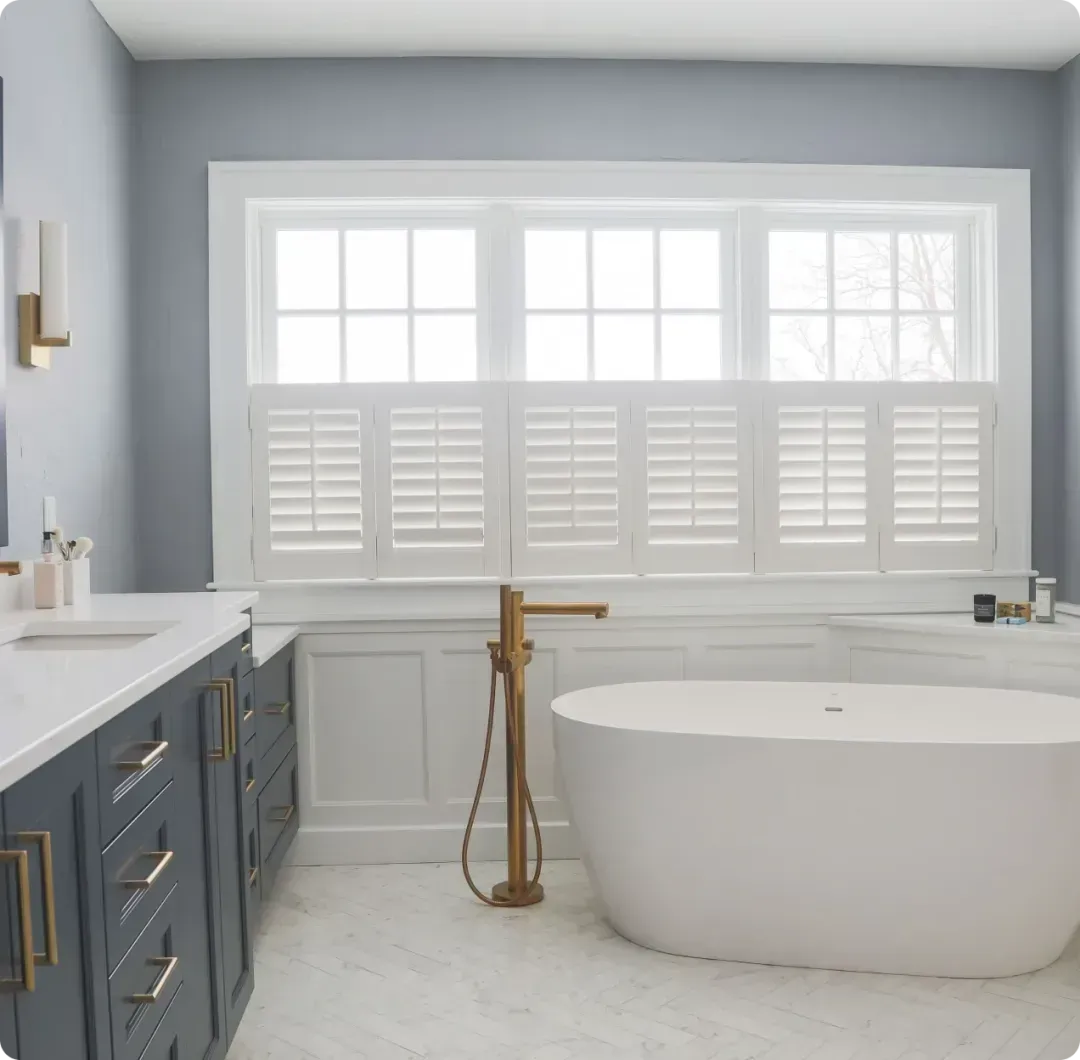 Modern bathroom with blue vanity, freestanding tub, brass fixtures, and bright window with shutters