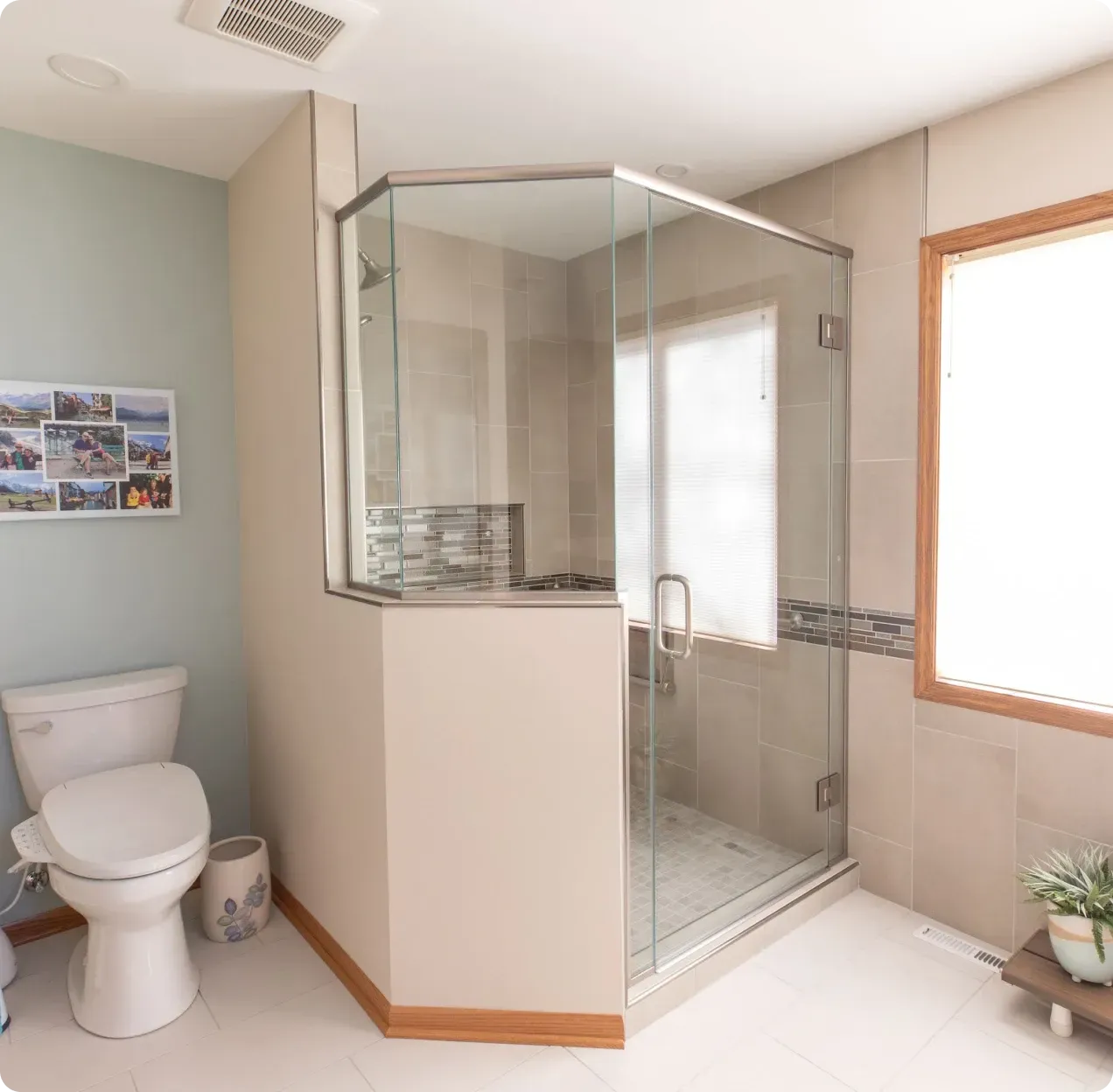 Bright accessible bathroom with toilet, glass shower, beige tile, and window light