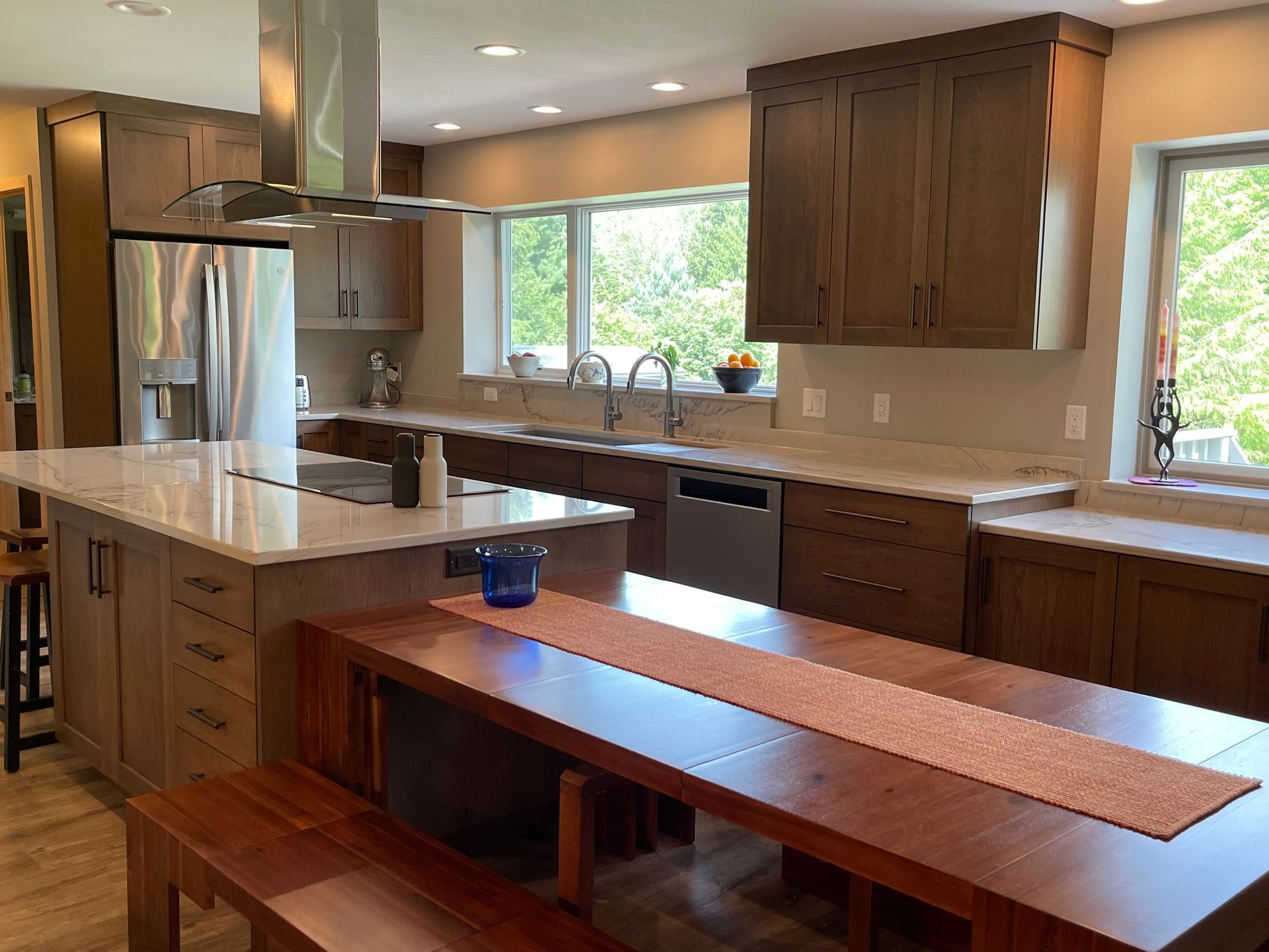 Modern kitchen with island, wooden table and benches, stainless steel appliances, and large windows.