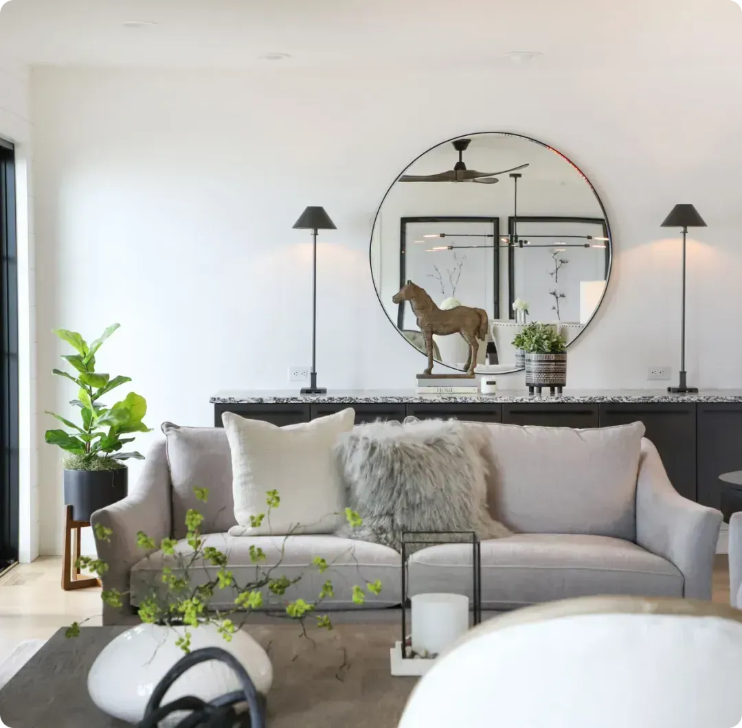 Bright living room with a gray sofa, round mirror, lamps, plants, and a cat on the console table
