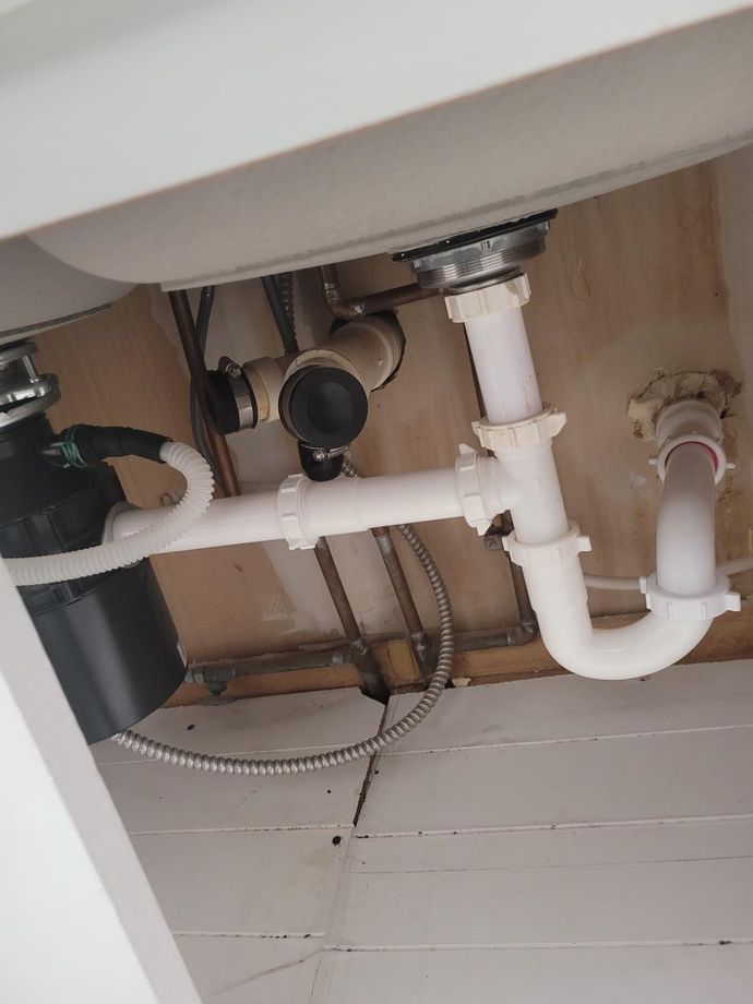 Under-sink plumbing with white PVC pipes, valve, and drain connections against a wood cabinet wall