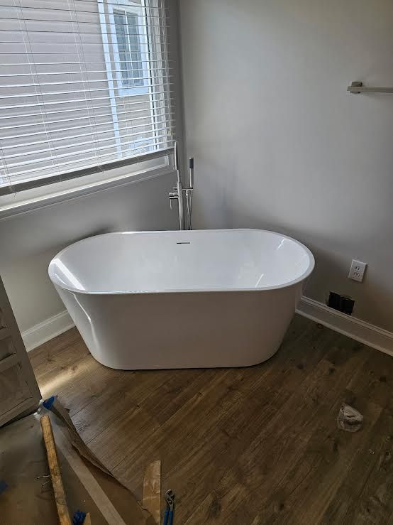 Freestanding white bathtub in a partially renovated room with wood flooring and a window blind.
