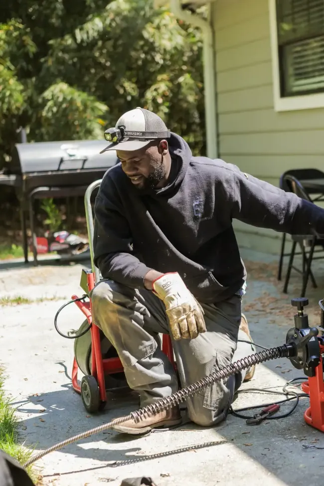 Worker kneeling outdoors with gloves and cap, using a power tool on a driveway near a house.