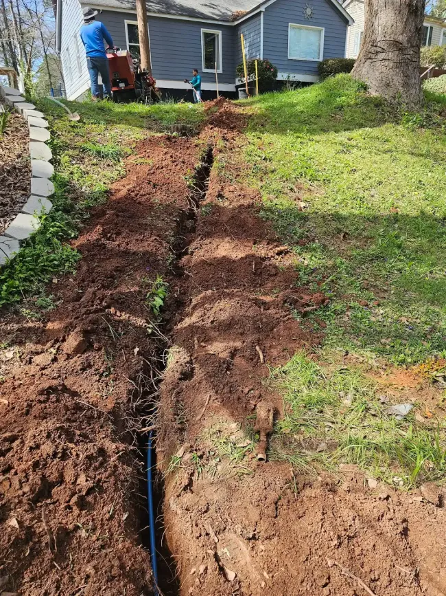 Fresh trench cut through a sloped yard to a blue house, exposing a black pipe line