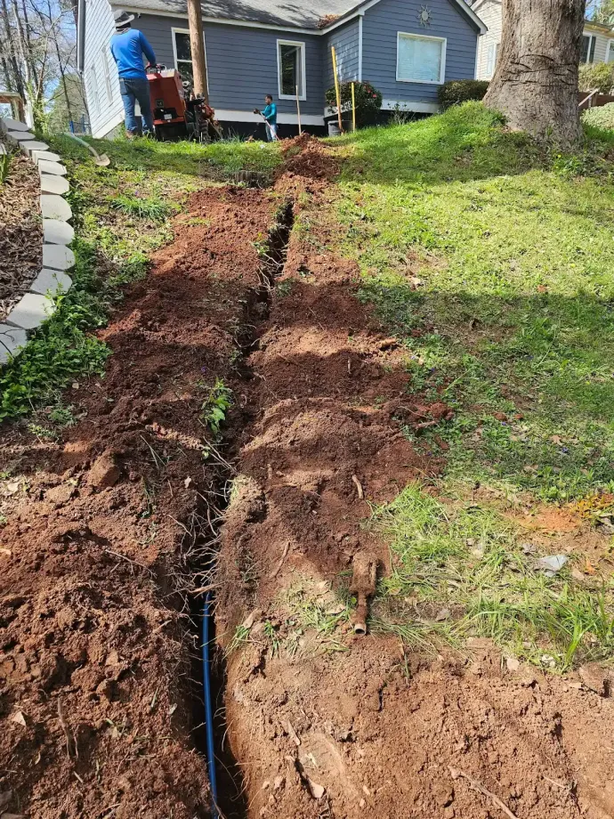 Fresh trench cut through a sloped yard to a blue house, exposing a black pipe line