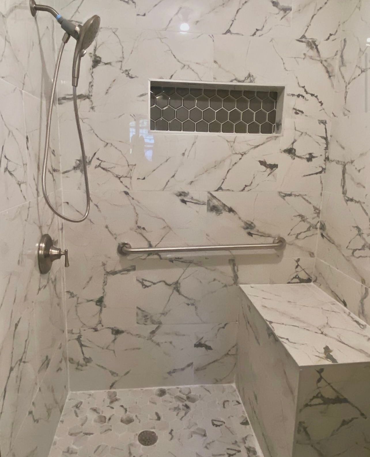 A white marble tiled shower with a built-in bench, silver grab bar, hand-held shower head, and a dark hexagonal niche.
