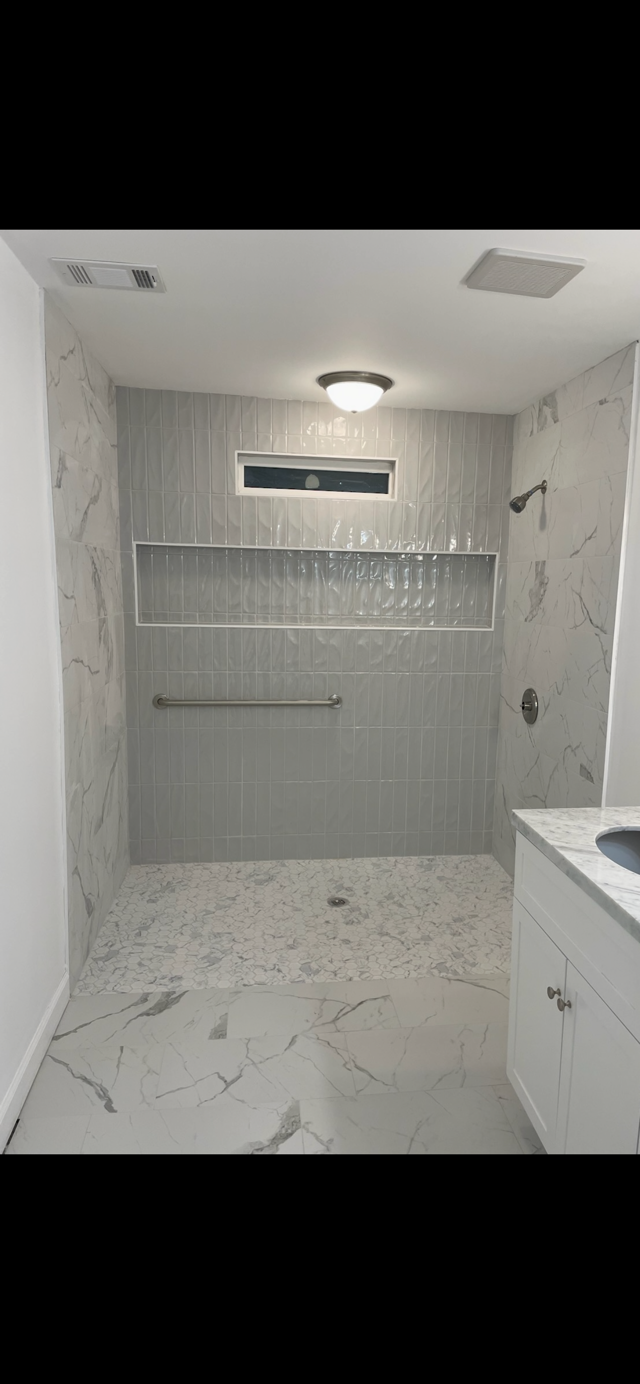 A modern shower with white marble walls, a gray-tiled accent wall, a long inset niche, a grab bar, and a marble floor.