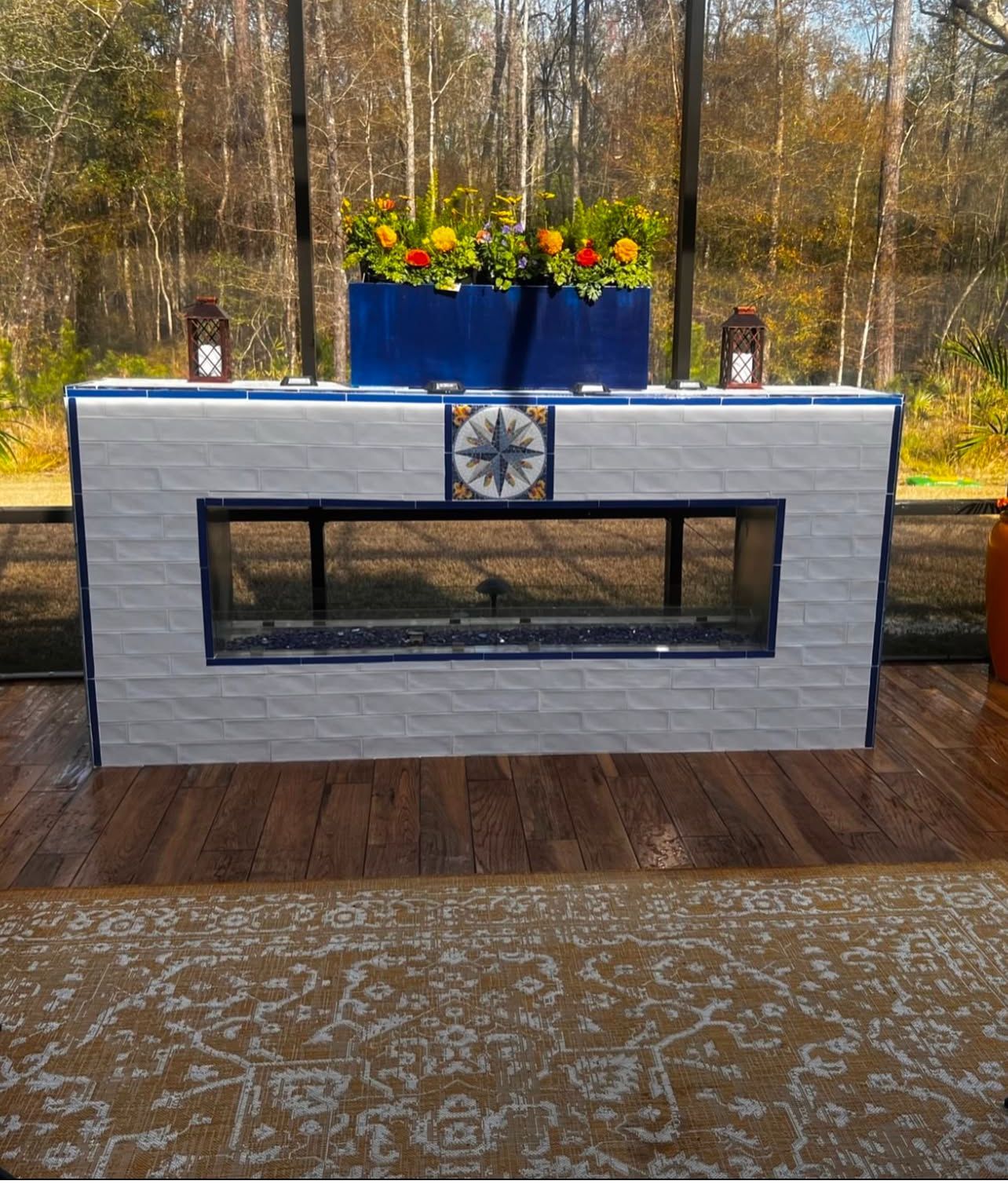A linear fireplace with white brick and blue trim, featuring a decorative compass tile and a blue planter with flowers.