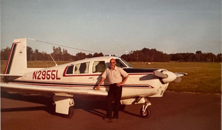 A person stands beside a white aircraft with red and blue stripes and the registration number N2955L on an airfield.