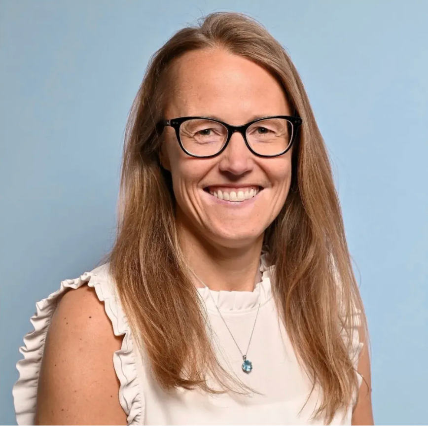 A person with long blonde hair wearing glasses and a white ruffled top, smiling against a solid blue background.