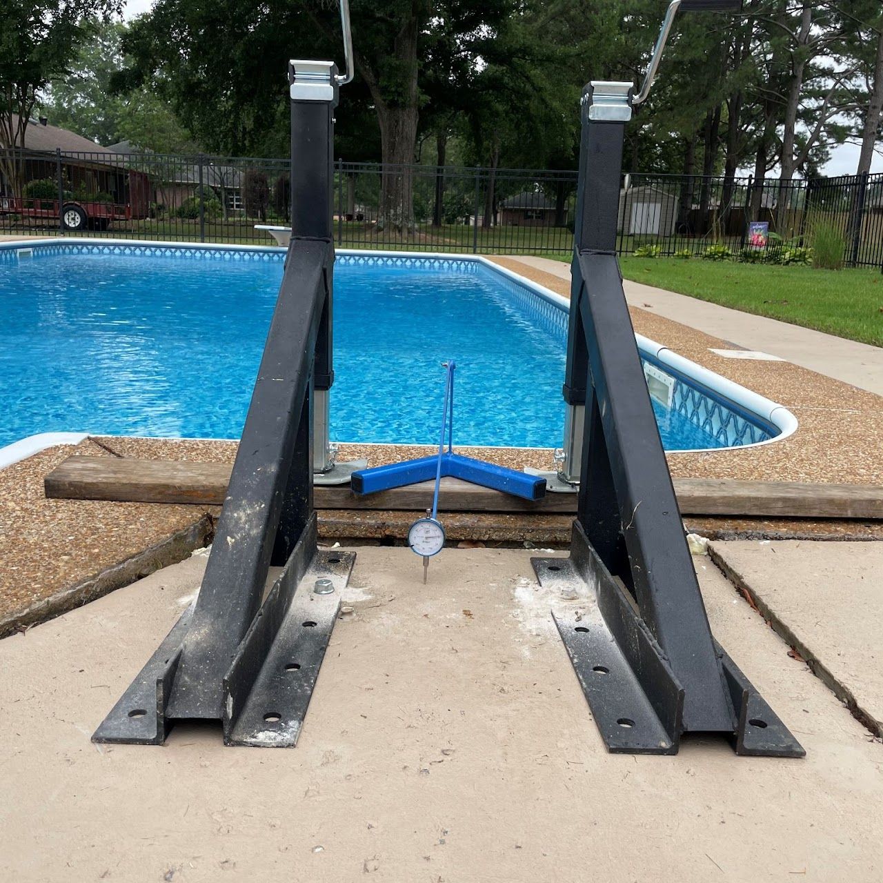 A dial gauge measures the gap between two black metal pool-ladder anchors mounted on a concrete pool deck.