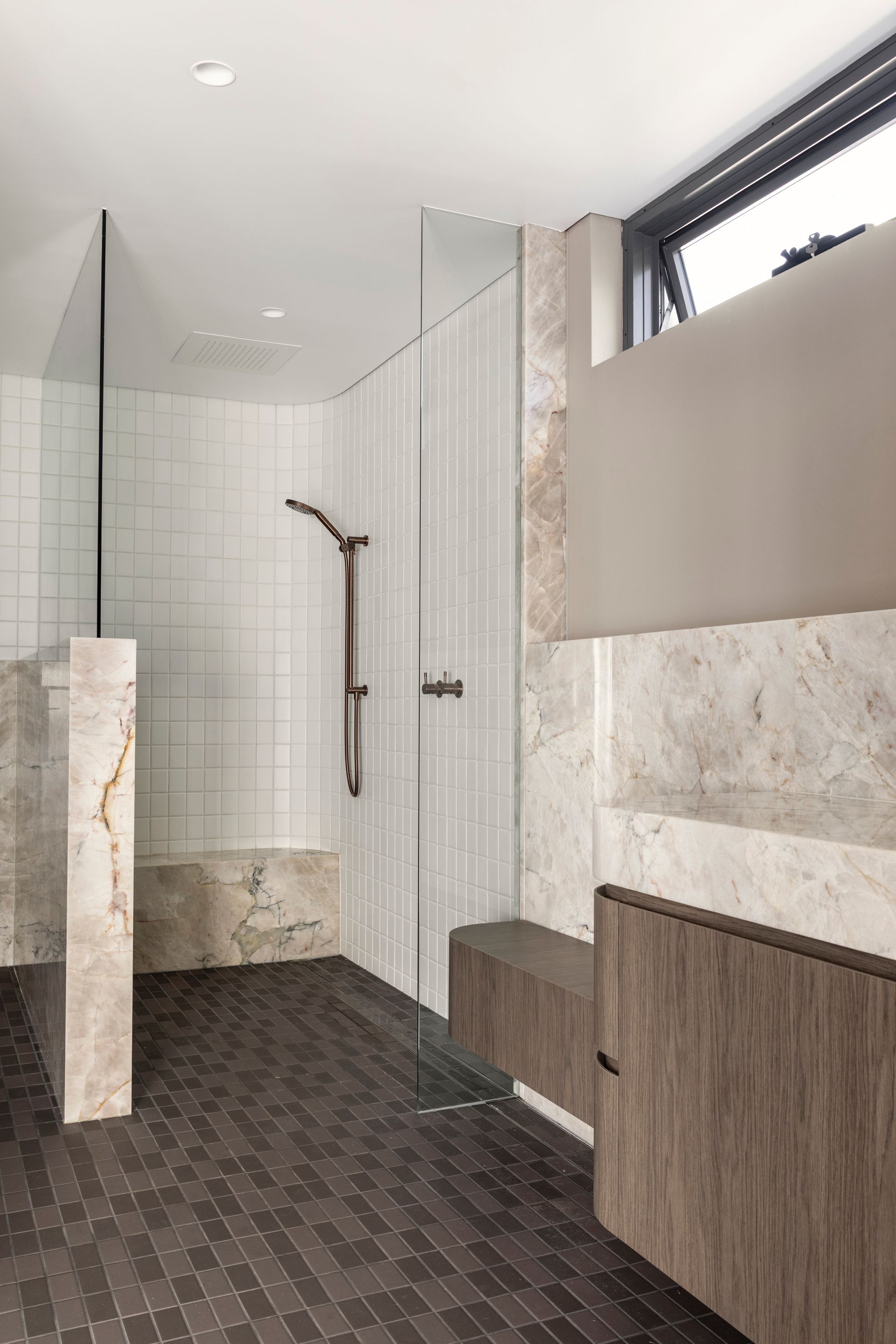 Modern bathroom with tiled shower, marble accents, wood cabinets, and a high window