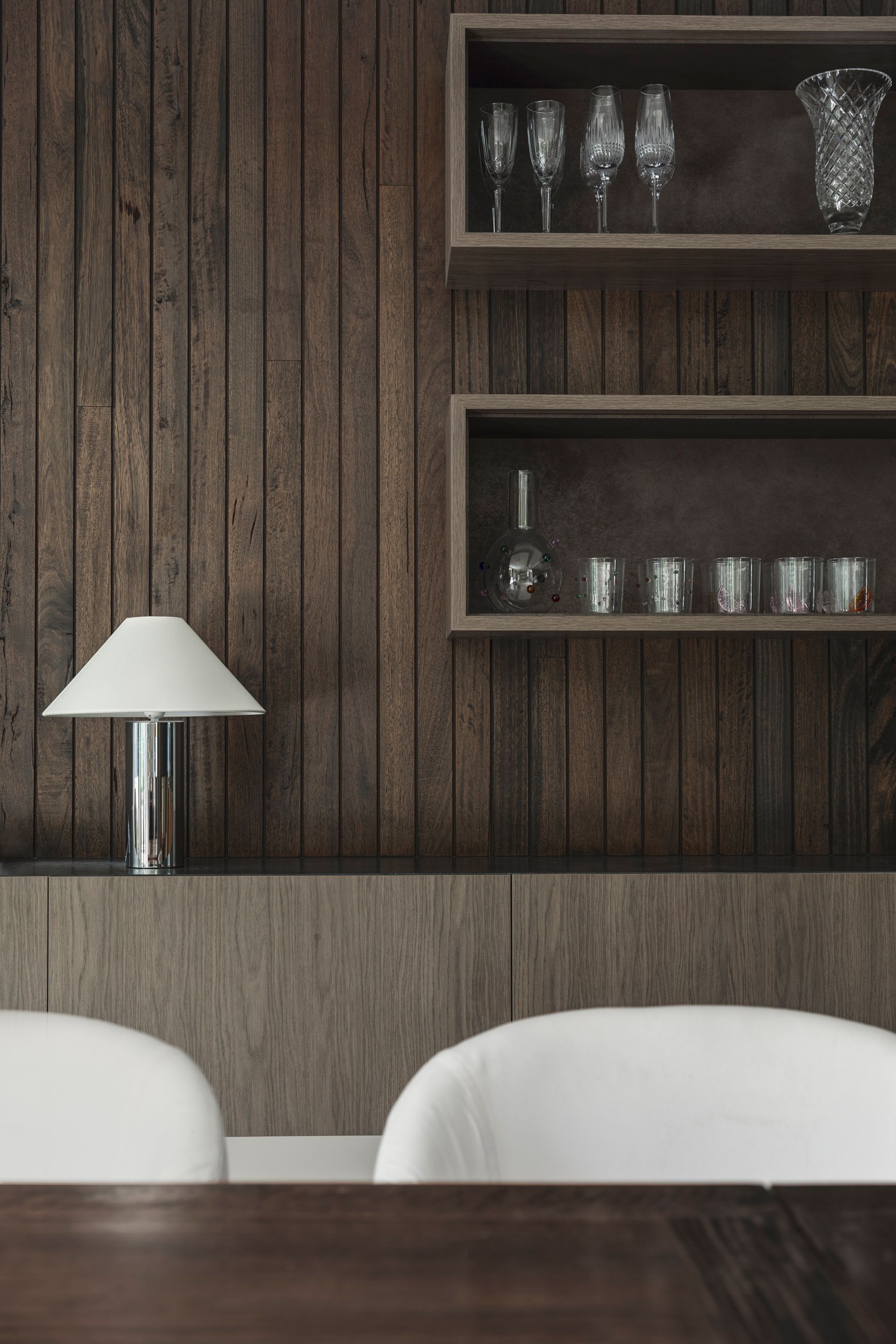 Modern dining room with wood-paneled wall, shelves of glassware, and white lamp above white chairs