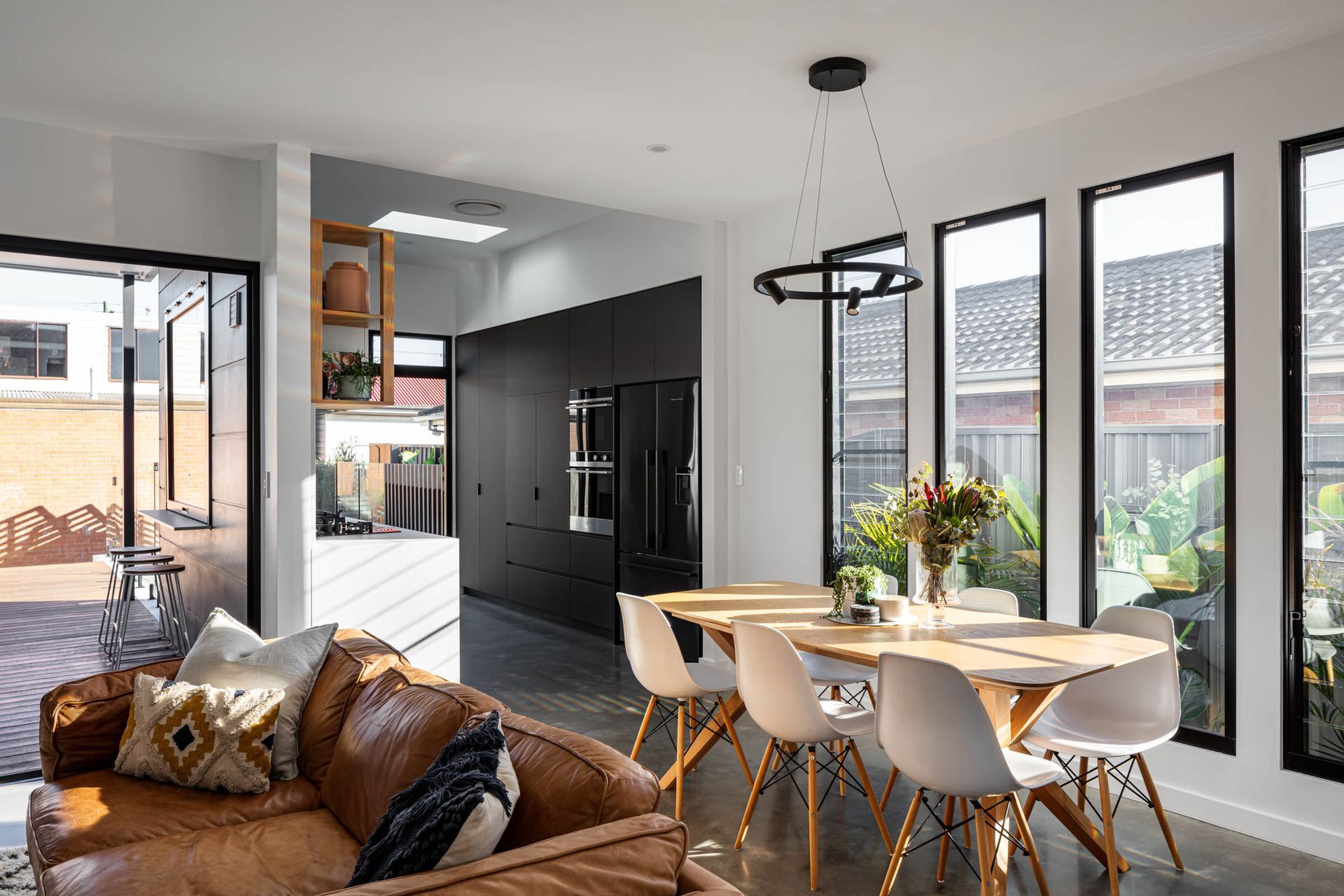 Open-plan living space with brown leather couch, dining table, black cabinetry, and tall windows.— Goode By Design in Unanderra, NSW