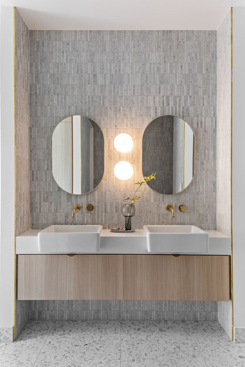 Double bathroom vanity with oval mirrors, light fixture, and light wood cabinet against a tiled wall. — Goode By Design in Unanderra, NSW