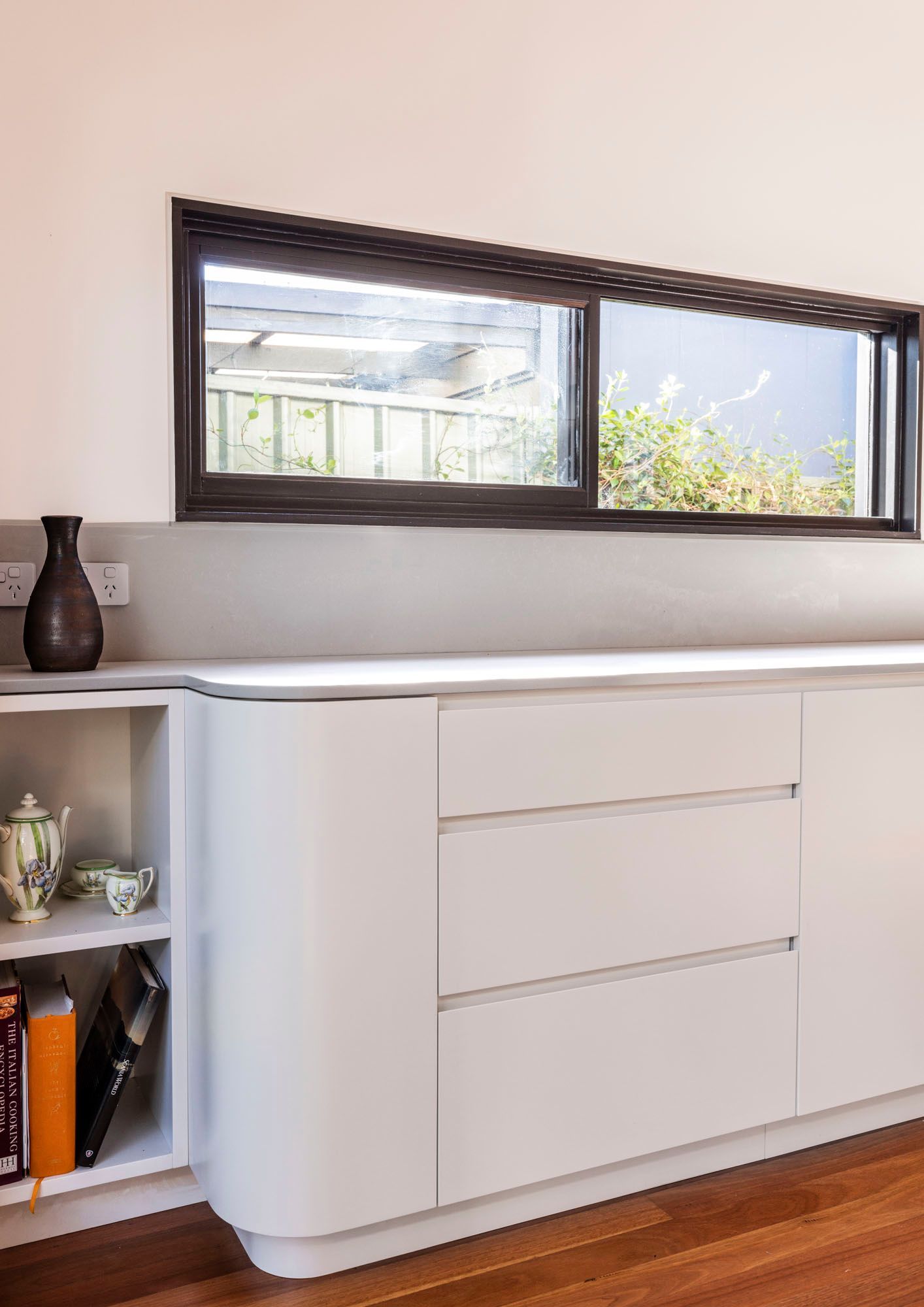 White cabinet with drawers, built-in bookshelf, and long window. A vase and tea set are visible. — Goode By Design in Unanderra, NSW