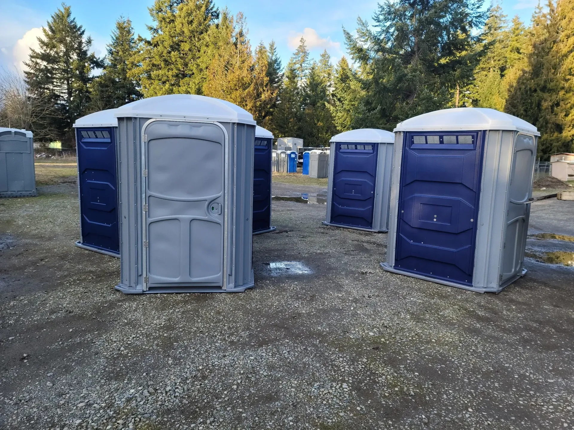Portable toilets on a gravel lot, with trees in the background.
