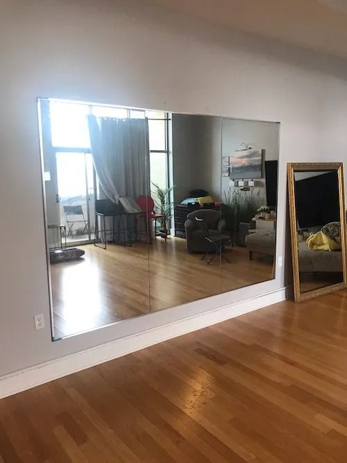 Large wall mirror reflects a bright room with hardwood floors, a window, and furniture.
