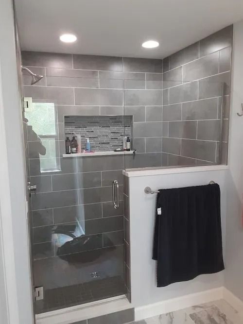 Gray-tiled shower with glass door and built-in shelf. A black towel hangs on the outside.