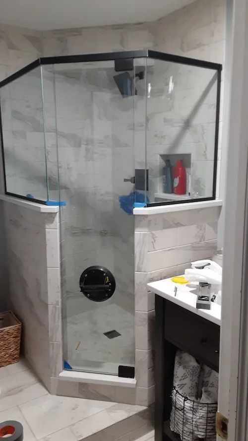 A newly tiled shower with a black-framed glass enclosure. A vanity and basket are visible.