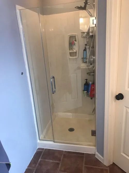 Shower stall with glass door, beige walls, various toiletries on shelves, and brown tile flooring.