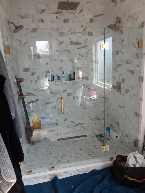 Modern shower with marble-like tile, glass doors, and gold fixtures. Two shower heads, small windows, and toiletries visible.