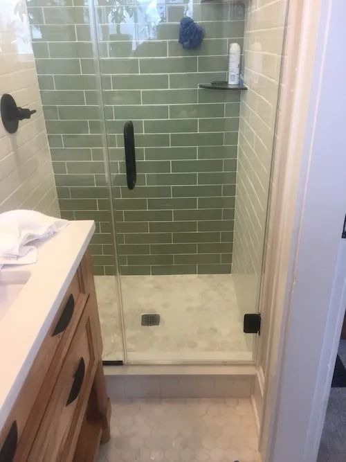Shower stall with green tile and glass door, next to a wooden vanity with black hardware.