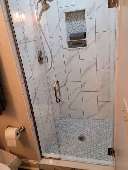 Modern shower with clear glass door, marble-look tile, built-in niche, and gray hexagon floor tiles.
