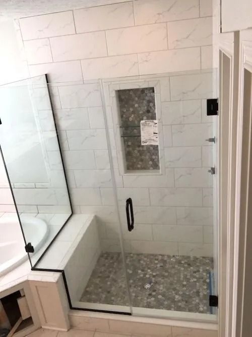 A newly renovated shower with white marble-like tiles and glass doors with black framing, featuring a built-in bench.