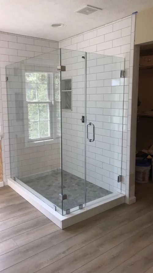 Glass shower enclosure with white tile walls and gray tile floor, next to a window and wood-look flooring.