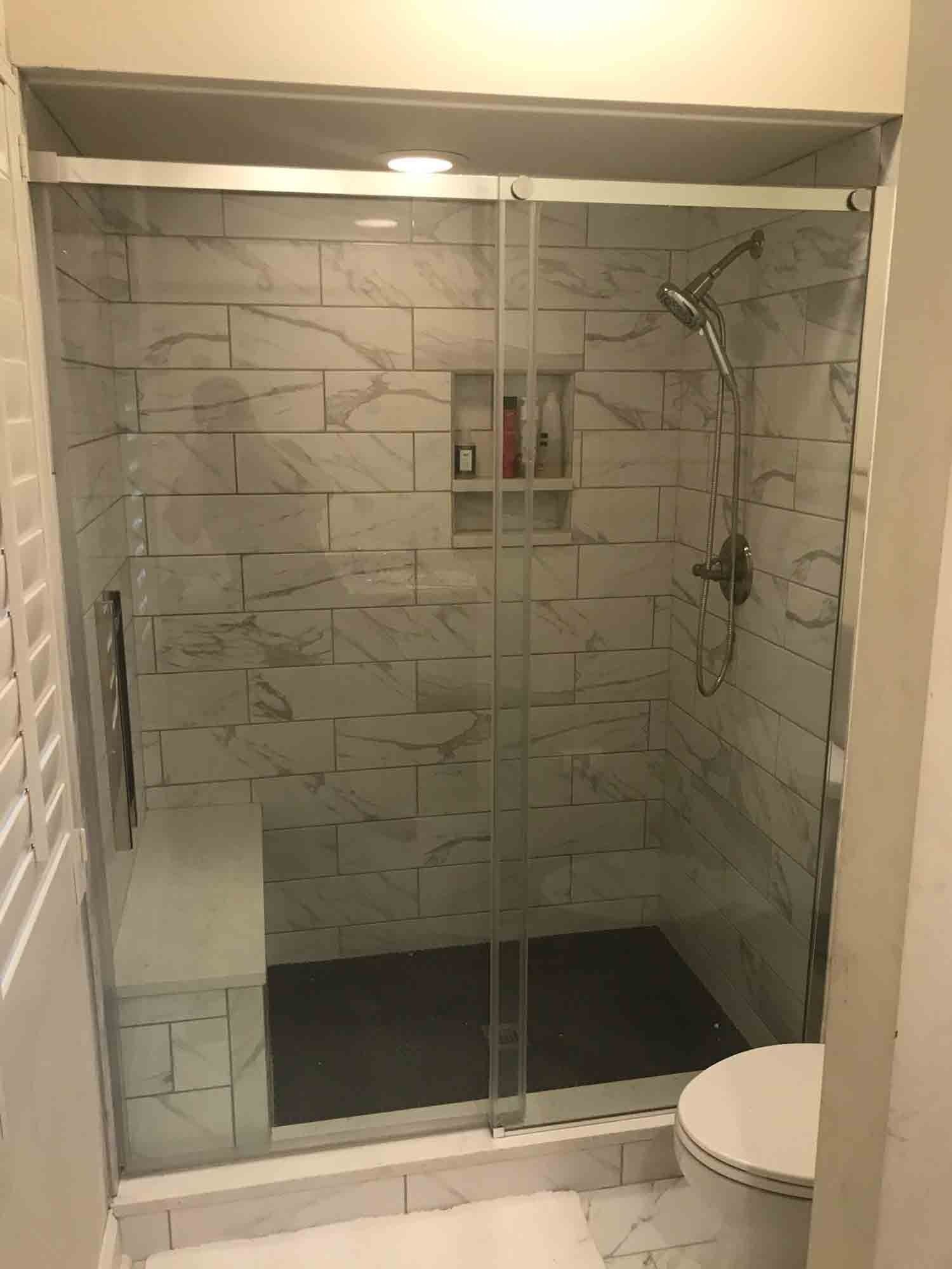 Shower stall with glass doors, white tiled walls, built-in bench, showerhead, and a toilet.
