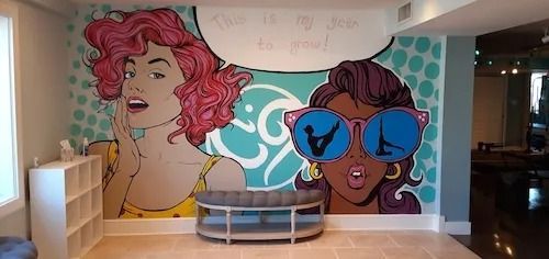 Colorful mural with pop art women in a room; one w/ pink hair and the other wearing sunglasses. A bench sits in front.