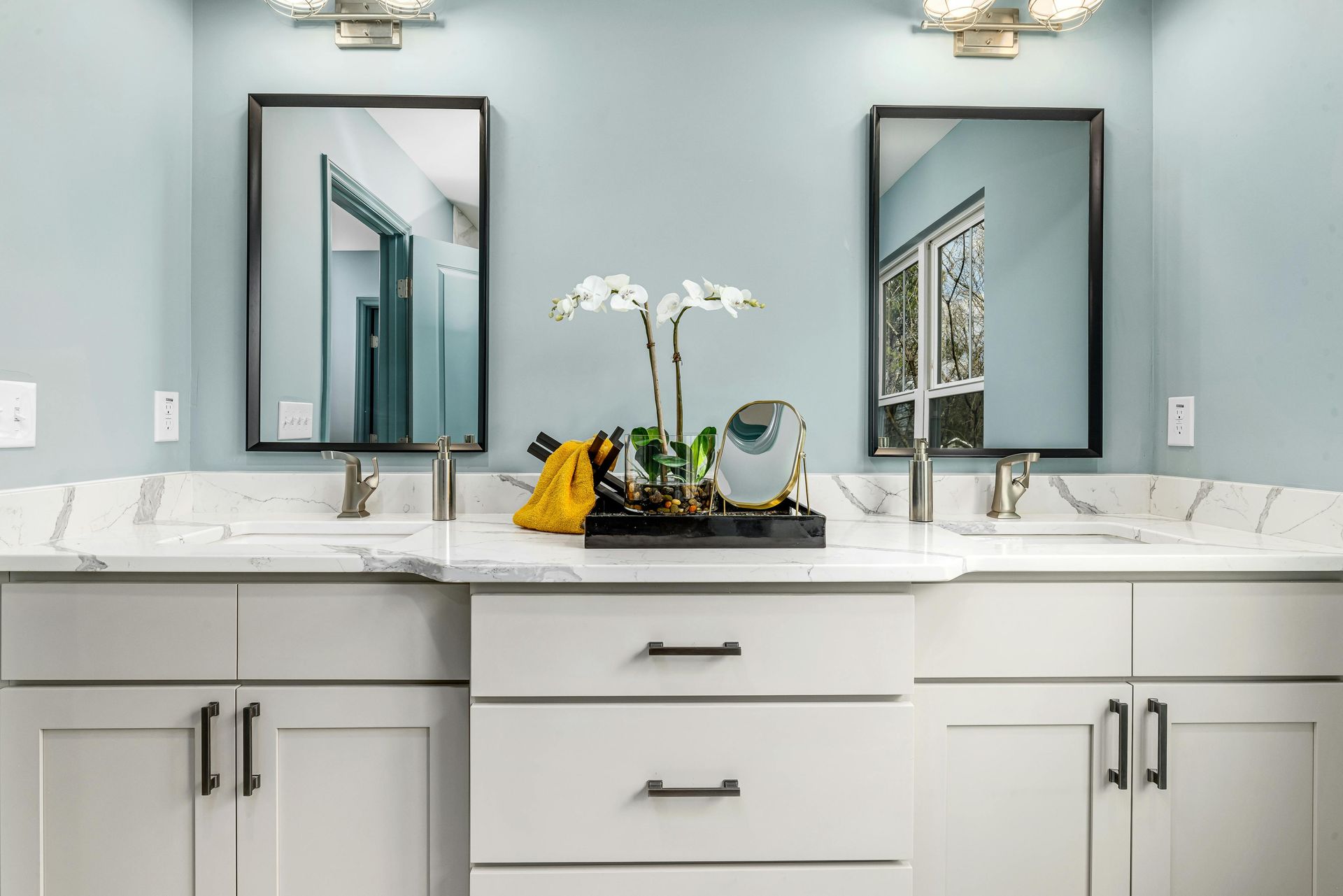 Double vanity bathroom with mirrors, white cabinets, and blue walls. Decor includes flowers and a decorative tray.