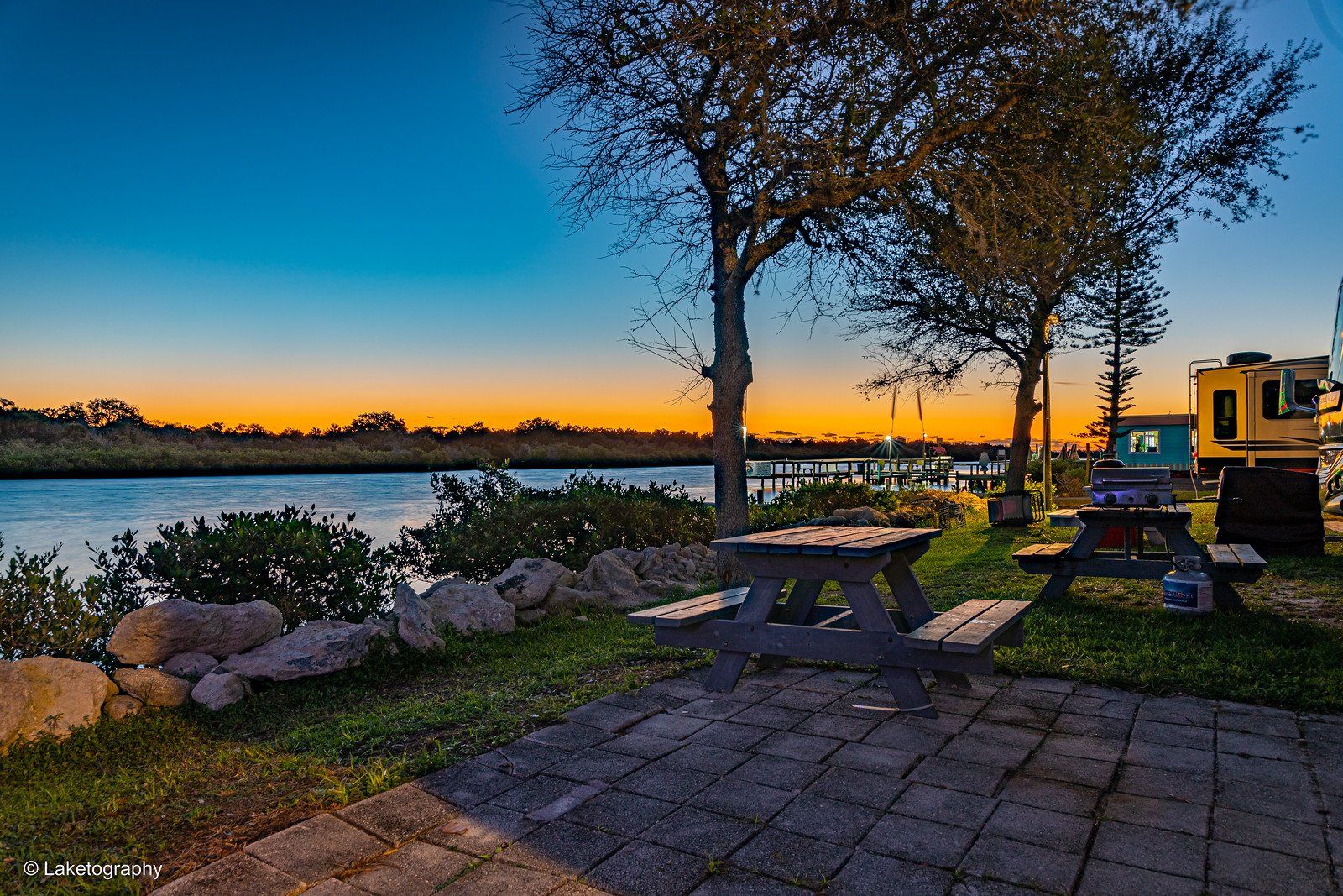 A picnic table is sitting next to a body of water at sunset.