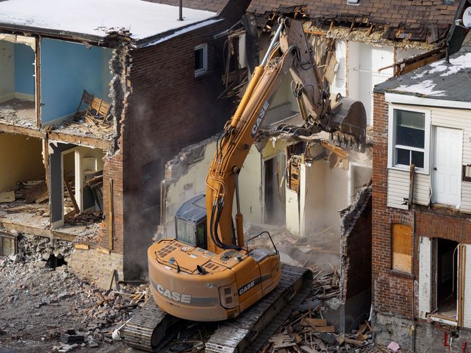 Yellow excavator demolishing a damaged house beside neighboring homes on a debris-filled lot
