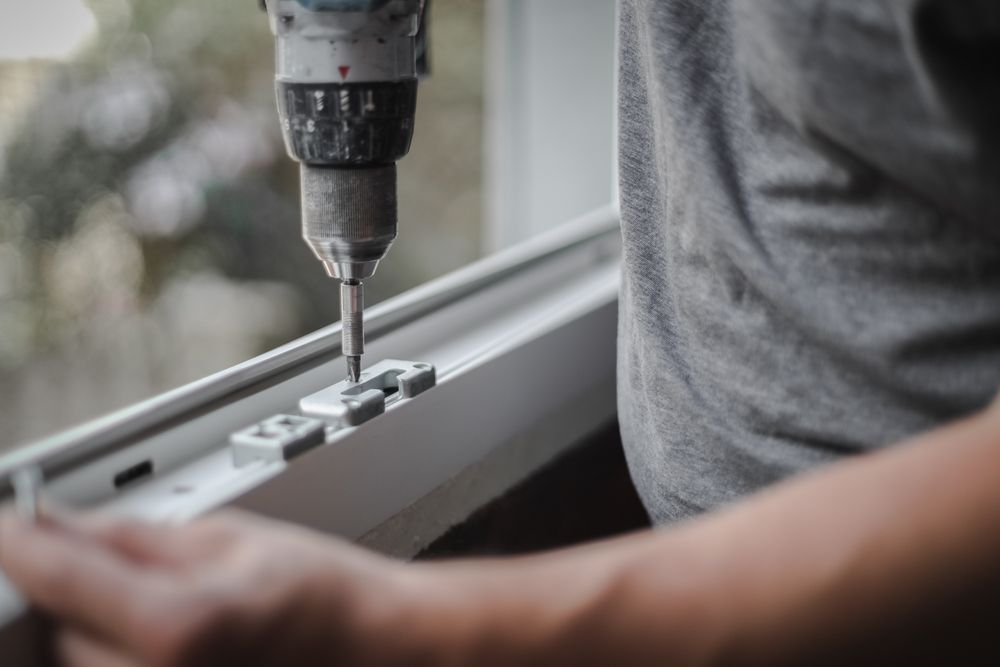 A person uses a power drill to install metal window hardware onto a white frame.