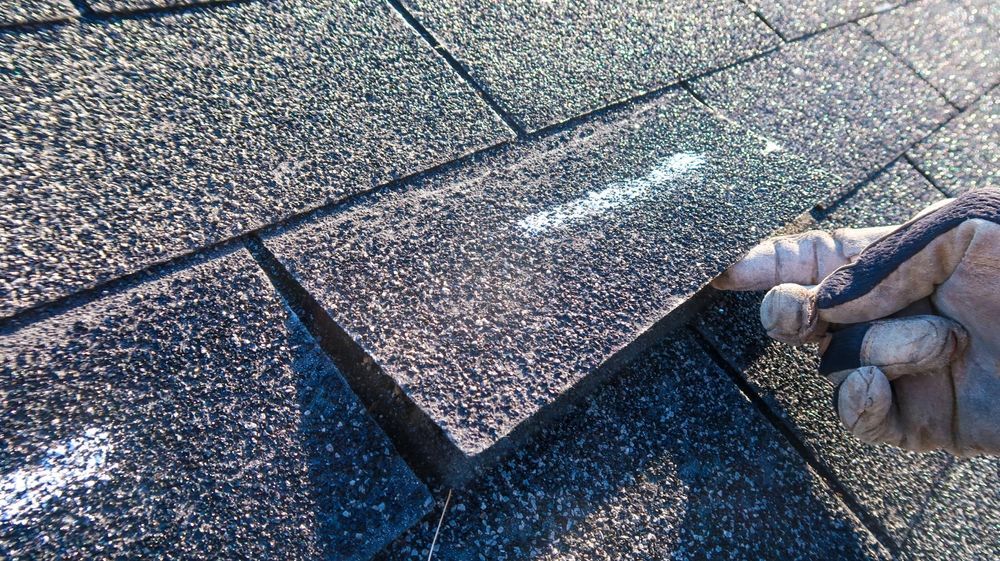A gloved hand lifts the corner of a dark asphalt roof shingle to reveal a white mark underneath on a sunny rooftop.