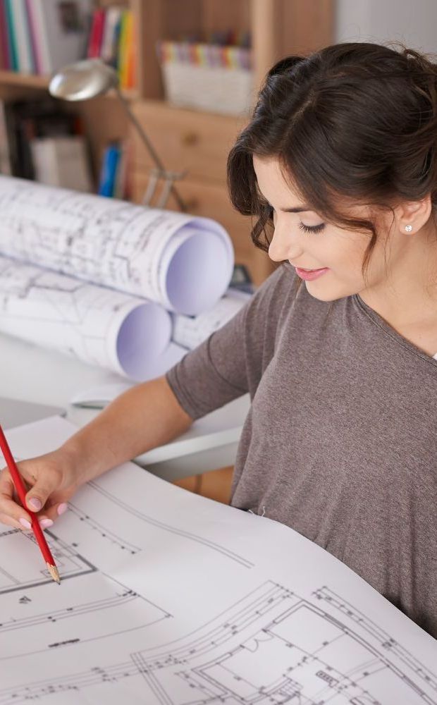 Una mujer está sentada en una mesa dibujando un plano con un lápiz rojo.