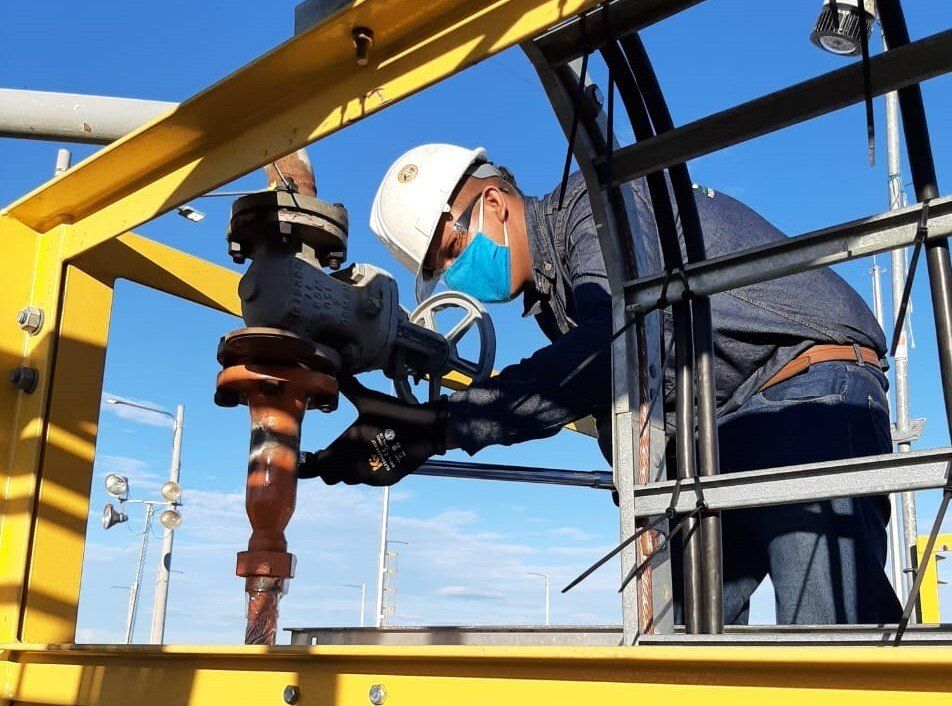 Trabajador con casco y mascarilla ajustando válvula en estructura industrial amarilla.