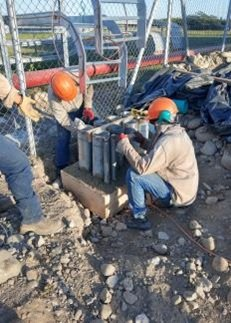 Obreros de la construcción con cascos de color naranja instalan postes de metal en una base de hormigón cerca de una valla.
