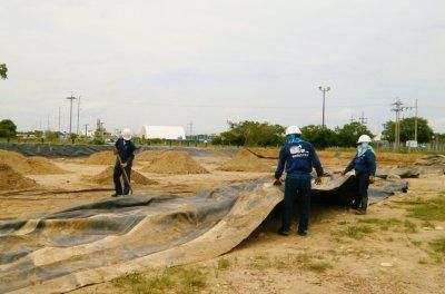 Tres trabajadores colocan una lona negra sobre una zona arenosa, con montones de arena al fondo.