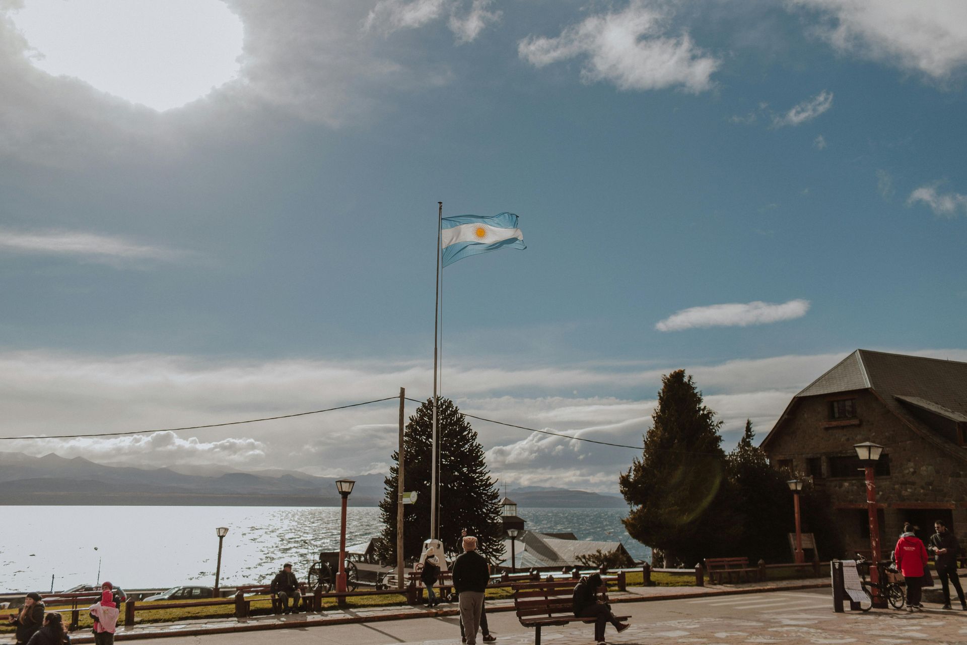 Un mástil sobre una plaza junto al lago con bancos, árboles y un edificio bajo un cielo nublado.