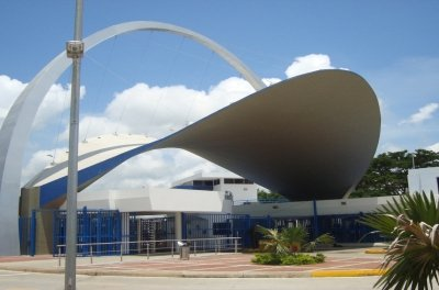 Entrada de edificio moderno con gran techo curvo de color gris y detalles en azul, bajo un arco blanco.