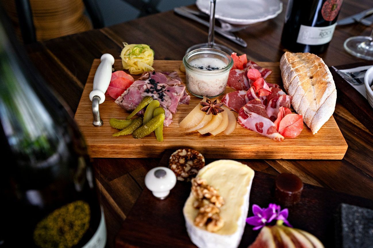 A wooden cutting board topped with meat , cheese , bread , pickles and a bottle of wine.