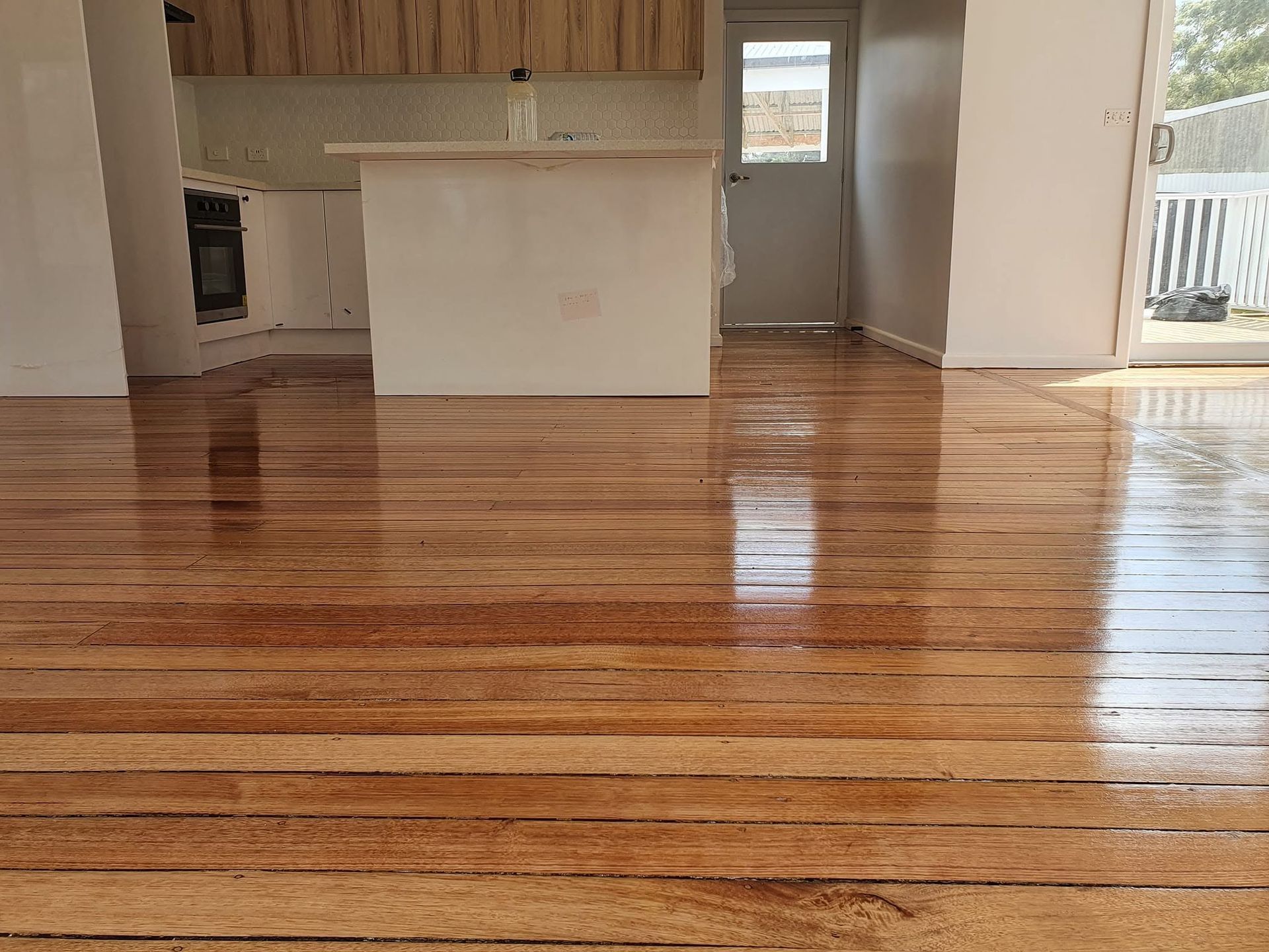 Empty room with glossy hardwood floors, kitchen area, and sliding glass door to a deck.